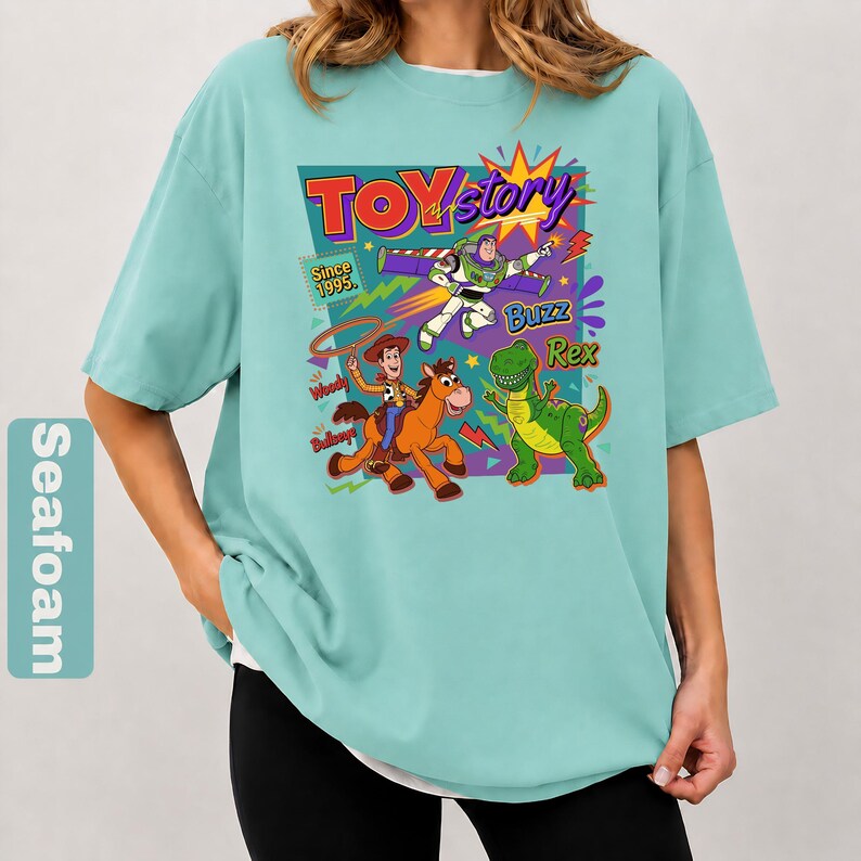 May include: Seafoam green t-shirt featuring a colorful graphic of Toy Story characters. The design includes Woody, Buzz Lightyear, Rex, and the Toy Story logo. The text "Since 1995" is also included in the design.