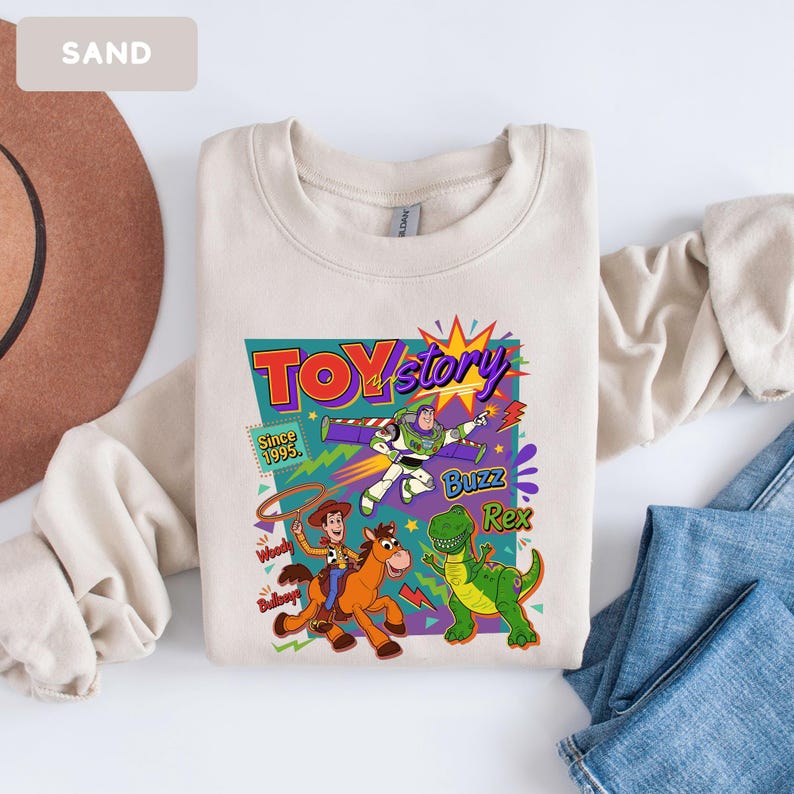 May include: Sand-colored sweatshirt featuring a colorful graphic of Toy Story characters. The design includes Buzz Lightyear, Woody, Rex, and the words "Toy Story" and "Since 1995". The sweatshirt is made of a soft material and is suitable for casual wear.