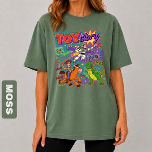 May include: Moss green t-shirt featuring a colorful graphic of Toy Story characters, including Woody, Buzz Lightyear, and Rex. The design includes the text "TOY STORY" and "Since 1995". The shirt has a relaxed fit.