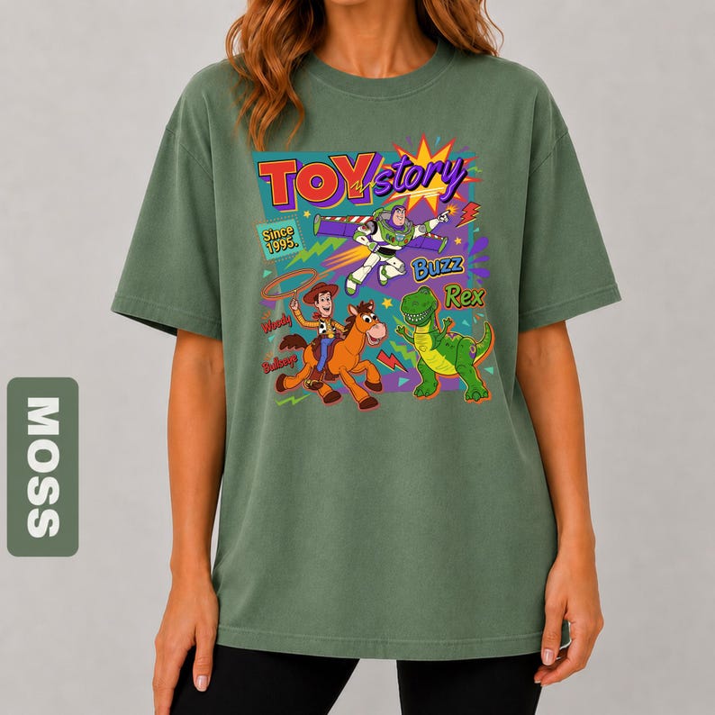 May include: Moss green t-shirt featuring a colorful graphic of Toy Story characters, including Woody, Buzz Lightyear, and Rex. The design includes the text "TOY STORY" and "Since 1995". The shirt has a relaxed fit.
