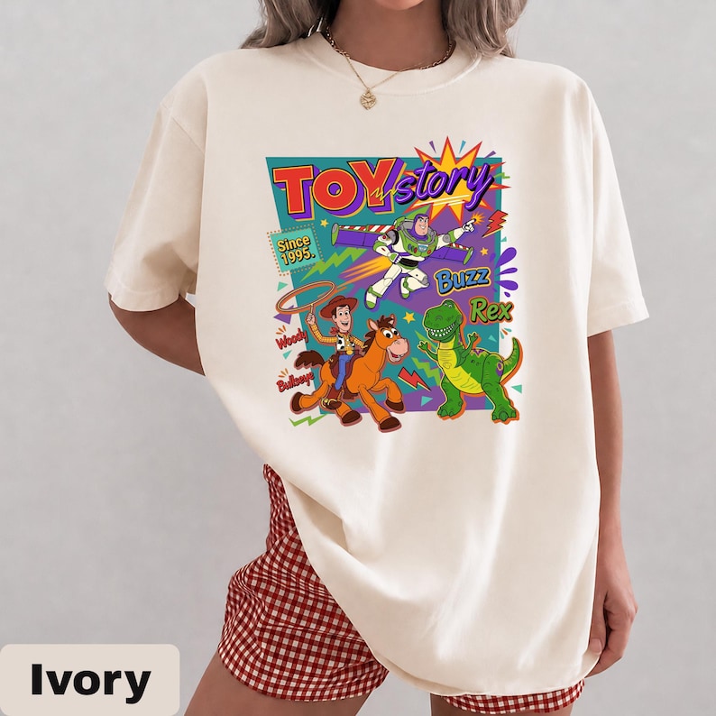May include: Ivory-colored t-shirt featuring a colorful graphic from the animated film "Toy Story." The design includes the film's title, characters Buzz Lightyear, Woody, Rex, and the text "Since 1995." The shirt has short sleeves and a crew neck.