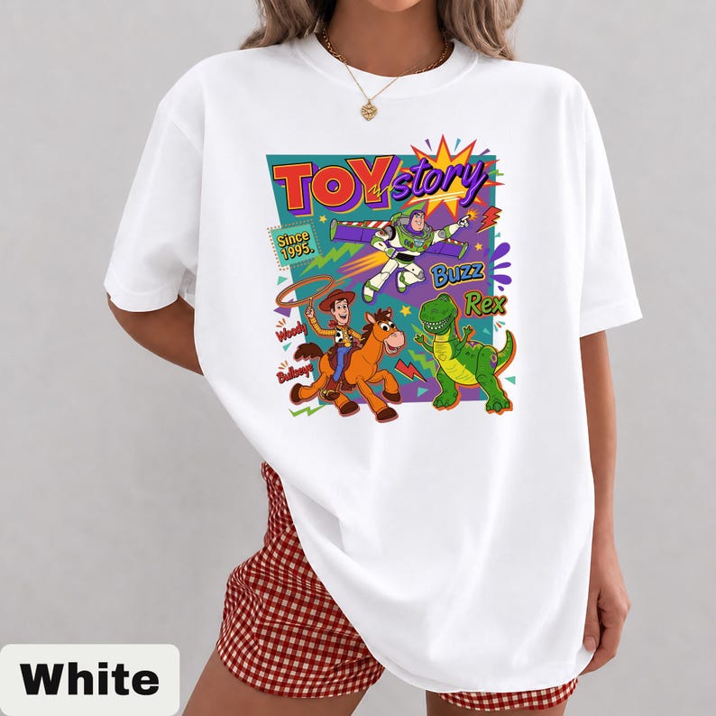 May include: White t-shirt featuring a colorful graphic of Toy Story characters, including Woody, Buzz Lightyear, and Rex. The design includes the text "Toy Story" and "Since 1995". The shirt is a casual, short-sleeved style.