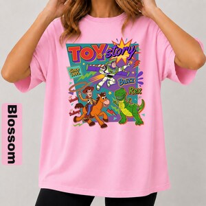 May include: A light pink t-shirt featuring a colorful graphic from the movie Toy Story. The design includes the words "Toy Story" and images of Woody, Buzz Lightyear, and Rex the dinosaur. The text "Since 1995" is also included.