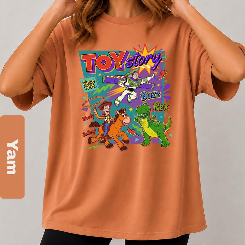 May include: An orange t-shirt featuring a colorful graphic from the movie Toy Story. The graphic includes the words "Toy Story" and images of Woody, Buzz Lightyear, and Rex the dinosaur. The text "Since 1995" is also included.