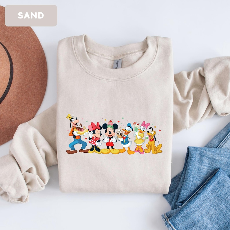 May include: A sand-coloured crewneck sweatshirt featuring a colourful graphic print of Goofy, Minnie Mouse, Mickey Mouse, Donald Duck, Daisy Duck, and Pluto holding various desserts, with the word SAND printed in the top left corner of the image.