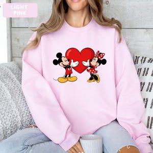 May include: A light pink crewneck sweatshirt featuring a graphic print of Mickey and Minnie Mouse holding a large red heart between them. The characters are depicted in their classic attire, with Mickey in red shorts and yellow shoes, and Minnie in a red polka-dot dress.