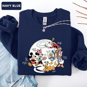 May include: Navy blue sweatshirt featuring a graphic of Mickey Mouse, Minnie Mouse, Donald Duck, Daisy Duck, Goofy, and Pluto. The characters are arranged around a cloud with the text "Mickey Mouse" and "Navy Blue". A silver necklace is draped on the sweatshirt.