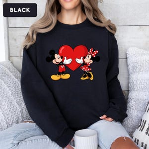May include: A black crewneck sweatshirt featuring a graphic print of Mickey and Minnie Mouse facing each other while holding a large red heart. The design includes Mickey in red shorts and yellow shoes, and Minnie in a red polka dot dress and bow.