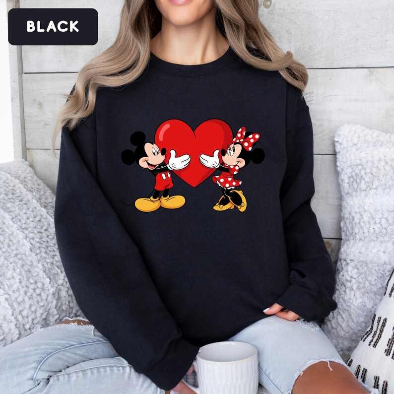 May include: A black crewneck sweatshirt featuring a graphic print of Mickey and Minnie Mouse facing each other while holding a large red heart. The design includes Mickey in red shorts and yellow shoes, and Minnie in a red polka dot dress and bow.