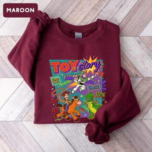 May include: Maroon sweatshirt featuring a colorful graphic from the movie Toy Story. The graphic includes the words "Toy Story" and the characters Buzz Lightyear, Woody, Bullseye, and Rex. The text "Since 1995" is also included.