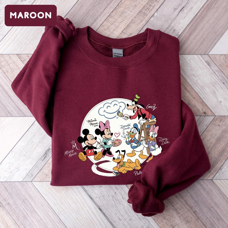 May include: Maroon sweatshirt featuring a graphic of Disney characters Mickey Mouse, Minnie Mouse, Donald Duck, Daisy Duck, Goofy, and Pluto. The characters are arranged around a cloud with a smiling face. The sweatshirt has long sleeves and a crew neck.