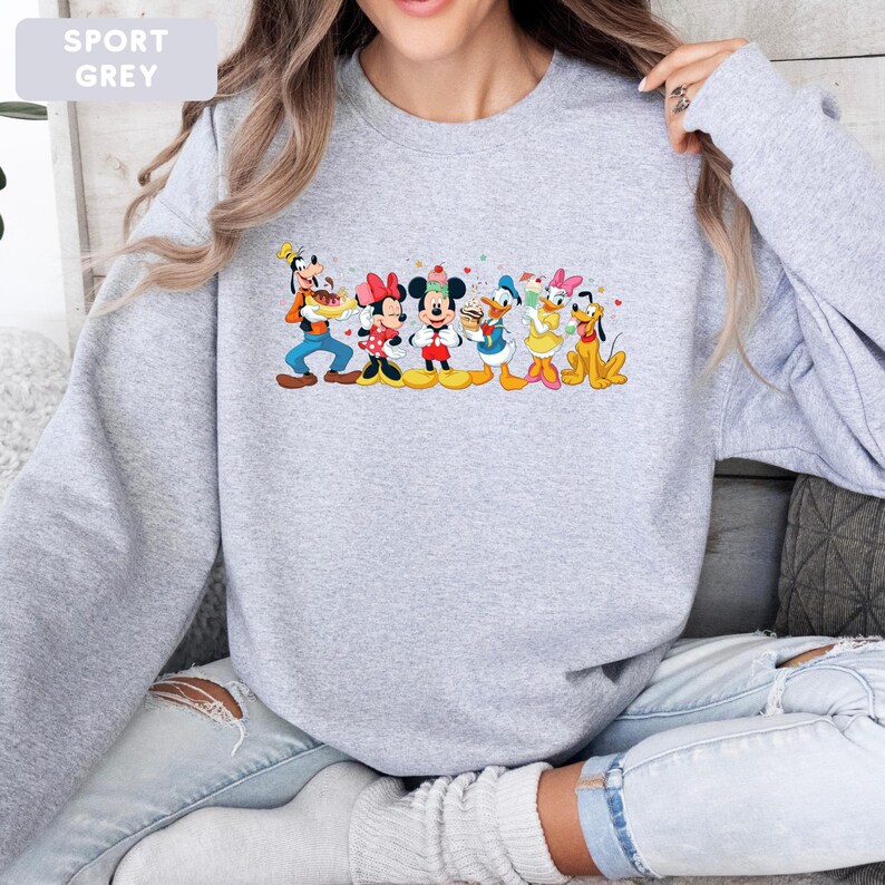 May include: A sport grey crewneck sweatshirt featuring a colourful graphic print of Disney characters including Goofy, Minnie Mouse, Mickey Mouse, Donald Duck, Daisy Duck, and Pluto. The characters are depicted standing together in a line.
