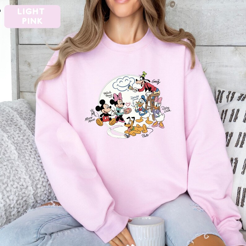 May include: Light pink sweatshirt featuring a Disney cartoon graphic. The graphic includes Mickey Mouse, Minnie Mouse, Donald Duck, Daisy Duck, Goofy, and Pluto. The words "Light Pink" are in a light pink rectangle in the upper left corner.