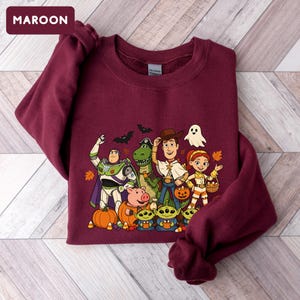 Puede incluir: Una sudadera de cuello redondo color granate con un estampado gr&aacute;fico de varios personajes animados vestidos con disfraces de Halloween, incluyendo un ranger espacial, un vaquero, una vaquera, un dinosaurio verde, un cerdo y tres peque&ntilde;os alien&iacute;genas verdes.