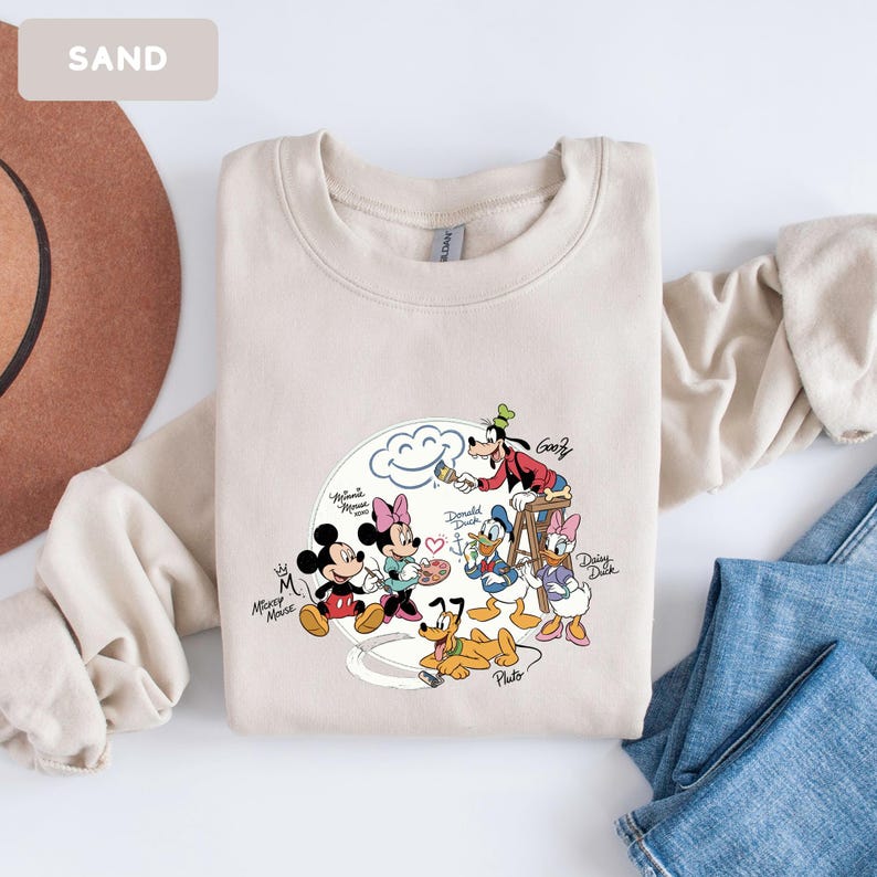 May include: Sand-colored sweatshirt featuring a graphic of Mickey Mouse, Minnie Mouse, Donald Duck, Daisy Duck, Goofy, and Pluto. The characters are depicted in a cartoon style, with a cloud and paintbrushes. The word "SAND" is in the upper left corner.