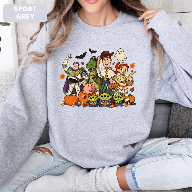 Puede incluir: Una sudadera gris sport de cuello redondo con un estampado gr&aacute;fico colorido de personajes de Toy Story disfrazados para Halloween. El dise&ntilde;o incluye a Buzz Lightyear, Rex, Woody, Jessie, Hamm y tres alien&iacute;genas rodeados de calabazas y hojas.