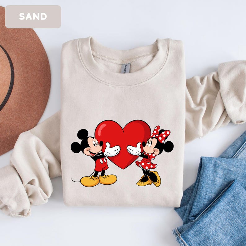 May include: A sand-colored crewneck sweatshirt featuring a graphic print of Mickey and Minnie Mouse holding a large red heart between them. The design shows Mickey in red shorts and yellow shoes, and Minnie in a red polka-dot dress and bow.