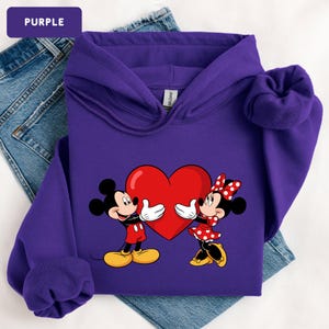 May include: A purple hooded sweatshirt featuring a graphic print of Mickey and Minnie Mouse holding a large red heart between them. The hoodie has long sleeves, a front pocket, and a drawstring hood. The text PURPLE is visible in a box.