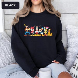 May include: A black long-sleeve sweatshirt featuring a colourful graphic print of Disney characters including Goofy, Minnie Mouse, Mickey Mouse, Donald Duck, Daisy Duck, and Pluto, each holding a dessert item, with small red hearts scattered above.