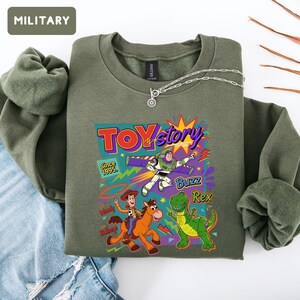 May include: An olive green sweatshirt featuring a colorful graphic of Toy Story characters, including Buzz Lightyear, Woody, and Rex. The graphic includes the text "Toy Story" and "Since 1995". A silver necklace is draped across the sweatshirt.