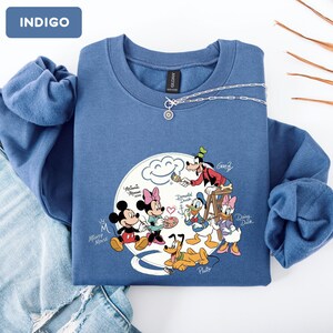 May include: Indigo blue sweatshirt featuring a Disney cartoon graphic. The graphic includes Mickey Mouse, Minnie Mouse, Donald Duck, Daisy Duck, Goofy, and Pluto. A silver necklace with a pendant is draped across the sweatshirt.