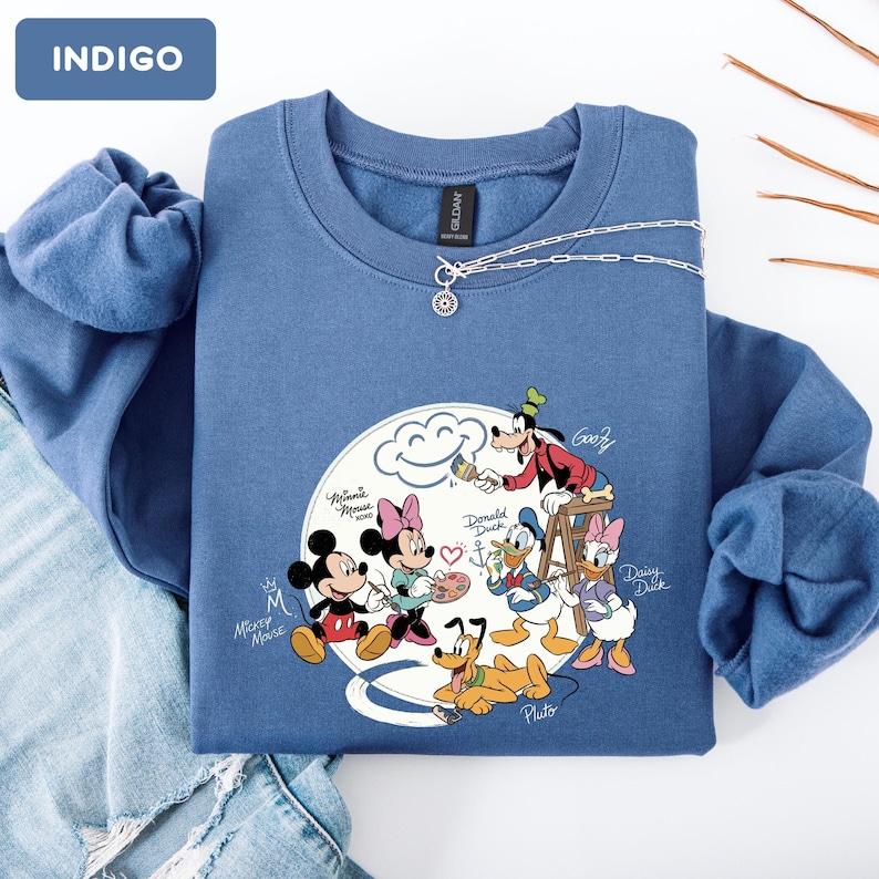 May include: Indigo blue sweatshirt featuring a Disney cartoon graphic. The graphic includes Mickey Mouse, Minnie Mouse, Donald Duck, Daisy Duck, Goofy, and Pluto. A silver necklace with a pendant is draped across the sweatshirt.