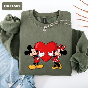 May include: An olive green crewneck sweatshirt featuring a graphic of Mickey and Minnie Mouse holding a large red heart between them. The sweatshirt has long sleeves with ribbed cuffs and a ribbed neckline. A silver chain necklace rests on the collar.