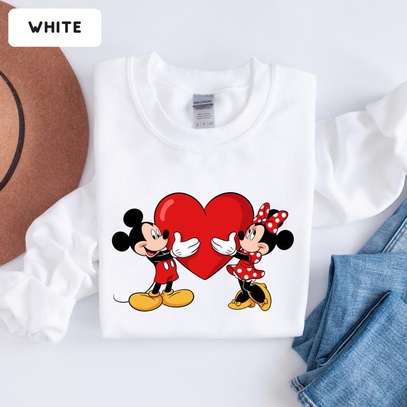 May include: A white long-sleeve sweatshirt featuring a graphic print of Mickey Mouse and Minnie Mouse holding a large red heart between them. The characters are depicted in their classic outfits with yellow shoes and white gloves on a plain white fabric.