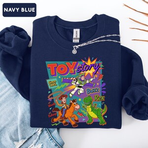 May include: Navy blue sweatshirt featuring a colorful graphic of Toy Story characters, including Buzz Lightyear, Woody, and Rex. The design includes the text "TOY Story" and "Since 1995." A silver necklace is draped across the sweatshirt.