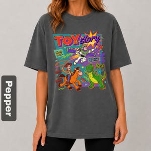 May include: Dark gray t-shirt featuring a colorful graphic from the movie Toy Story. The design includes the words "Toy Story" and images of Woody, Buzz Lightyear, and Rex the dinosaur. The text "Since 1995" is also included.