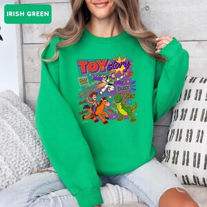 May include: Irish green sweatshirt featuring a colorful graphic from the movie Toy Story. The graphic includes the text "Toy Story Since 1995" and images of Woody, Buzz Lightyear, and Rex the dinosaur. The sweatshirt is a solid color.