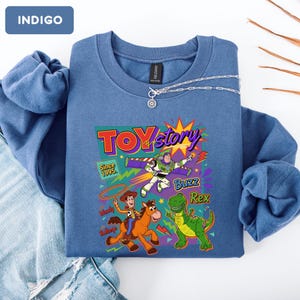 May include: Indigo blue sweatshirt featuring a colorful "Toy Story" graphic with Buzz Lightyear, Woody, and Rex. The graphic includes the text "Toy Story" and "Since 1995". A silver necklace with a pendant is draped on the sweatshirt.