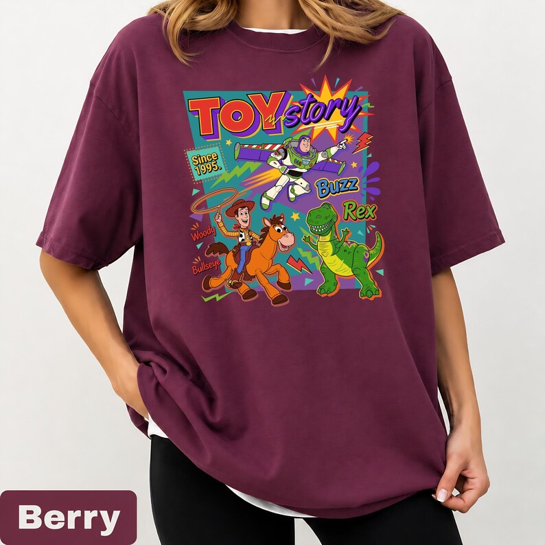 May include: Berry-colored t-shirt featuring a colorful graphic of Toy Story characters. The design includes Buzz Lightyear, Woody, Rex, and the text "TOY Story" and "Since 1995". The shirt has a relaxed fit and a white undershirt peeking out.