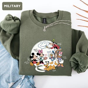 May include: Olive green sweatshirt with a crew neck and long sleeves. The sweatshirt features a graphic of Disney characters, including Mickey Mouse, Minnie Mouse, Donald Duck, and Goofy. A silver necklace with a pendant is on the sweatshirt. The word "MILITARY" is in a box in the upper left corner.