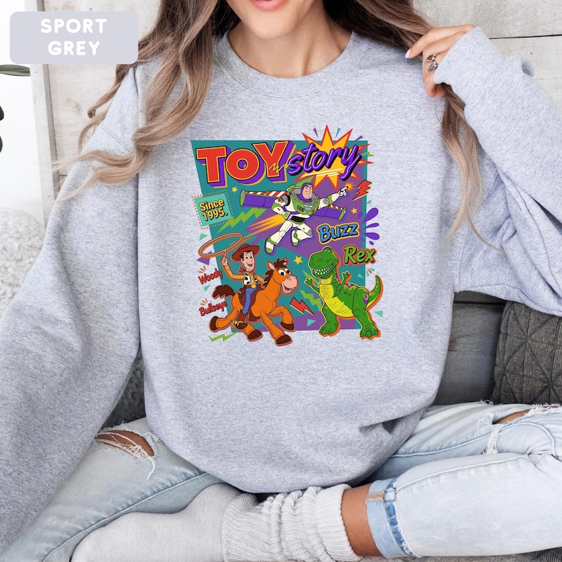 May include: Sport grey sweatshirt featuring a colorful Toy Story graphic. The design includes Woody, Buzz Lightyear, Rex, and the Toy Story logo with the text "Since 1995". The sweatshirt is a comfortable, casual style.