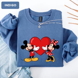 May include: An indigo crewneck sweatshirt featuring a graphic of Mickey and Minnie Mouse holding a large red heart. The sweatshirt has a round neckline and long sleeves. A small tag labeled INDIGO is visible in the top left corner of the image.