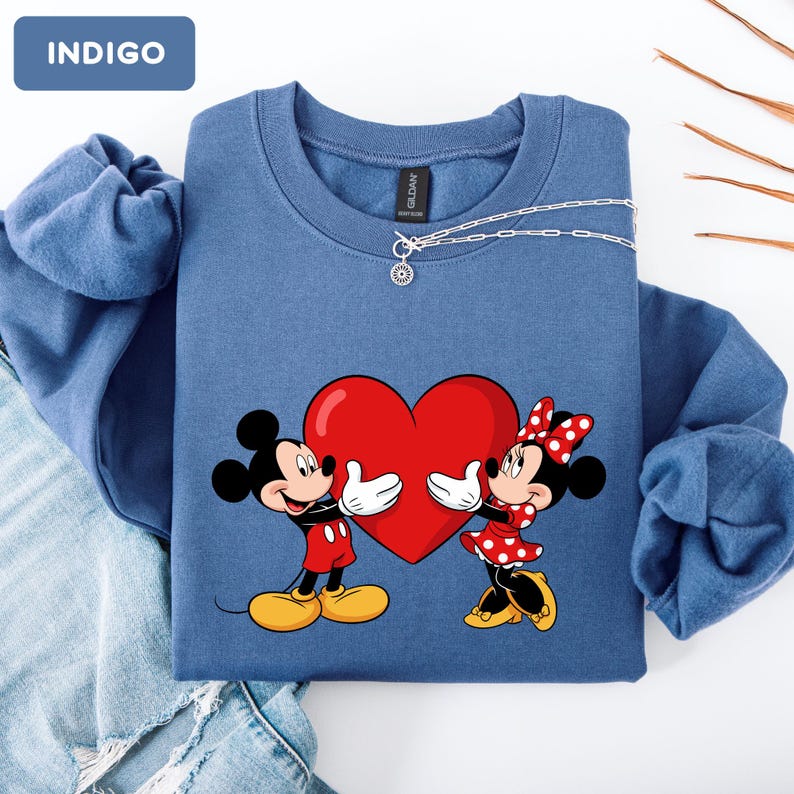 May include: An indigo crewneck sweatshirt featuring a graphic of Mickey and Minnie Mouse holding a large red heart. The sweatshirt has a round neckline and long sleeves. A small tag labeled INDIGO is visible in the top left corner of the image.