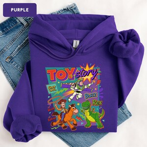 May include: A purple hooded sweatshirt featuring a graphic from the movie Toy Story. The graphic includes the words "Toy Story" and the characters Buzz Lightyear, Woody, Bullseye, and Rex. The sweatshirt is displayed on top of blue jeans.