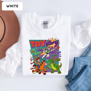 May include: White sweatshirt featuring a colorful graphic from the movie Toy Story. The graphic includes the text "Toy Story" and images of Woody, Buzz Lightyear, and Rex the dinosaur. The sweatshirt has a crew neck.