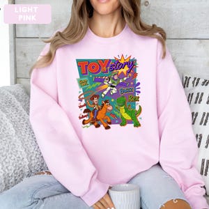 May include: Light pink sweatshirt featuring a colorful graphic from the movie "Toy Story." The design includes the words "Toy Story," "Buzz," "Rex," and "Woody." The sweatshirt has long sleeves and a crew neck.