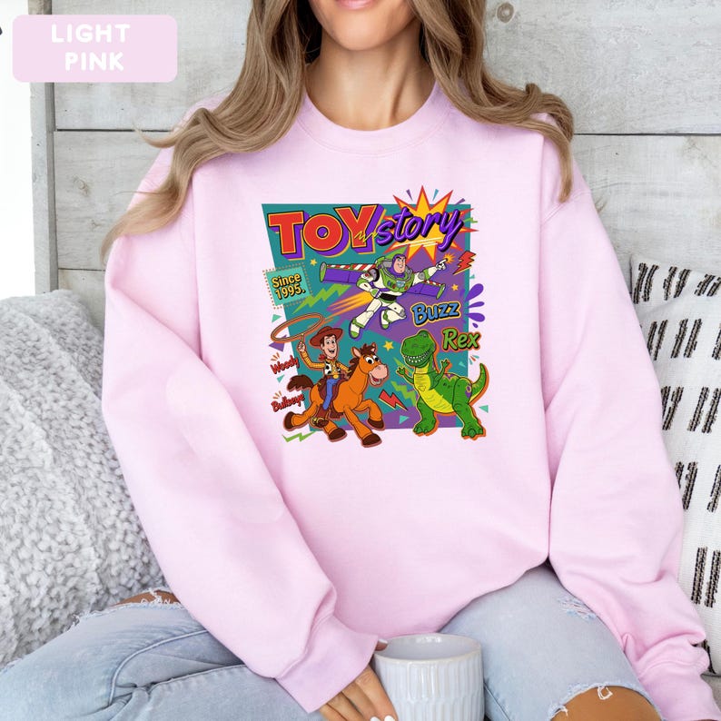 May include: Light pink sweatshirt featuring a colorful graphic from the movie "Toy Story." The design includes the words "Toy Story," "Buzz," "Rex," and "Woody." The sweatshirt has long sleeves and a crew neck.
