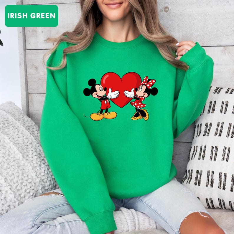 May include: An Irish green crewneck sweatshirt featuring a graphic print of Mickey and Minnie Mouse holding a large red heart between them. The design shows Mickey in red shorts and yellow shoes, and Minnie in a red polka dot dress and bow.