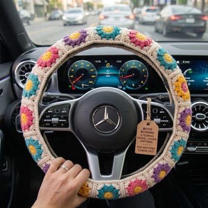 May include: A crocheted steering wheel cover with a floral pattern in shades of pink, yellow, purple, and teal. The cover is beige and has a tag that reads "Boho Blooms Steering Wheel Cover Pattern".