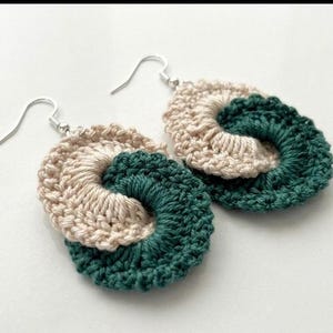 May include: Handmade crochet earrings in a spiral design. The earrings feature a combination of beige and teal yarn, with silver-colored ear wires. The earrings are a unique accessory for everyday wear.