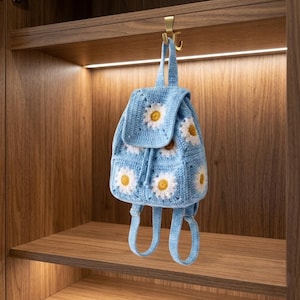 May include: A light blue crochet backpack featuring white and yellow daisy flower patterns. The backpack has a flap closure and adjustable straps. The bag is suspended from a gold hook inside a wooden cabinet.