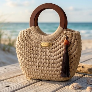 Peut inclure: Un sac à main beige en crochet avec une poignée en bois et une pompon décoratif. Le sac comporte une plaque signalétique dorée qui dit "Whisper". Le sac est posé sur une surface en bois près de la plage.