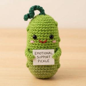 May include: A handmade, green crochet pickle with a smiling face, holding a sign that reads "EMOTIONAL SUPPORT PICKLE." The pickle has a green stem and is set against a light wooden background. A cute and whimsical item.