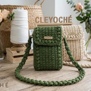 May include: Handmade green crochet crossbody bag with a matching strap. A beige crochet bag is also visible. The bags are displayed on a wooden table with the brand name "CLEYOCHÉ" in the foreground. Flowers and decorative items are in the background.