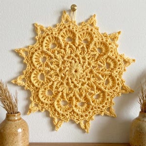 Könnte beinhalten: Ein gelber gehäkelter Wandbehang im Sonnenstrahl-Design hängt an einer weißen Wand. Zwei kleine Keramikvasen mit getrockneten Gräsern sind darunter platziert. Das Häkelstück hat einen Durchmesser von etwa 50 cm.