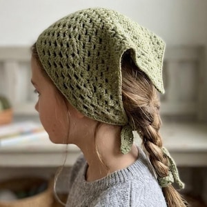 May include: A crocheted headscarf in a light olive green color. The head covering is square-shaped and tied at the nape of the neck. The crochet pattern has an open, lacy design. The scarf is worn over a braid.